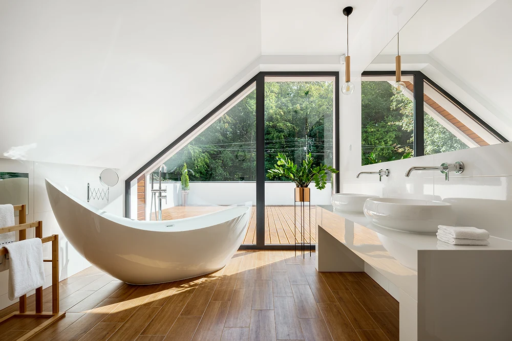 Ein modernes Badezimmer mit einer weißen freistehenden Badewanne neben großen dreieckigen Fenstern einer Immobilie in Münster. Auf der rechten Seite befinden sich zwei Waschbecken und eine hölzerne Arbeitsplatte. Das Sonnenlicht erhellt den Raum durch die Fenster und gibt den Blick auf das Grün draußen frei.