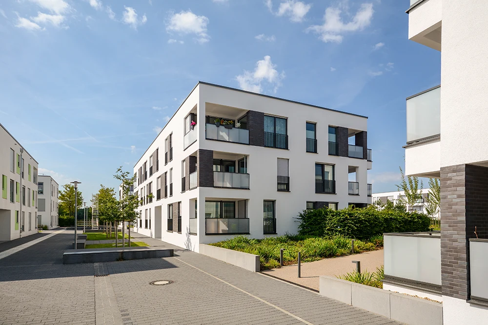 Moderne Wohnanlage mit weißen Gebäuden, großen Fenstern und Balkonen in Münster. Der Innenhof bietet eine gepflegte Grünanlage und einen gepflasterten Gehweg unter blauem Himmel. Bauträger ist Rosenke Immobilien aus Münster.