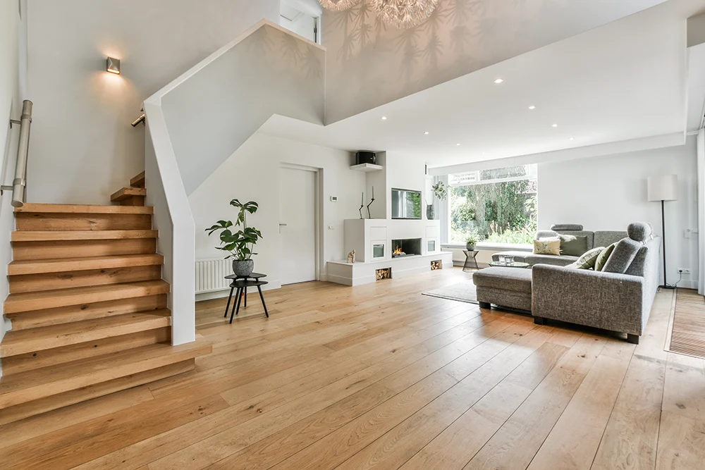 Geräumiges Wohnzimmer mit Holzfußboden, einem grauen Sektionssofa und einem großen Fenster, das natürliches Licht hereinlässt. Zur minimalistischen Einrichtung gehören eine Topfpflanze, ein an der Wand montierter Fernseher und eine weiße Treppe, die nach oben führt. Das Objekt wurde durch Rosenke Immobilien in Münster vermittelt.