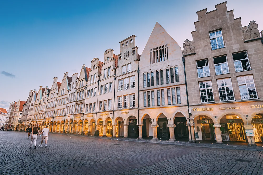 Eine malerische Ansicht des Prinzipalmarkts in Münster mit hohen, schmalen, giebelständigen Gebäuden. Die Architektur weist Bögen und Steinfassaden auf. Auf der kopfsteingepflasterten Straße gehen Menschen spazieren, und die Gebäude werden von Straßenlaternen warm beleuchtet.