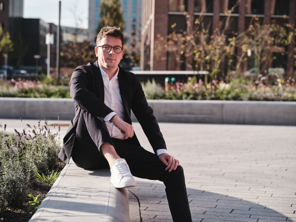 Nico Rosenke in einem schwarzen Anzug und weißen Turnschuhen sitzt lässig auf einem Steinsockel in städtischer Umgebung in Münster, umgeben von Grünflächen. Im Hintergrund sind moderne Gebäude und ein klarer Himmel zu sehen.